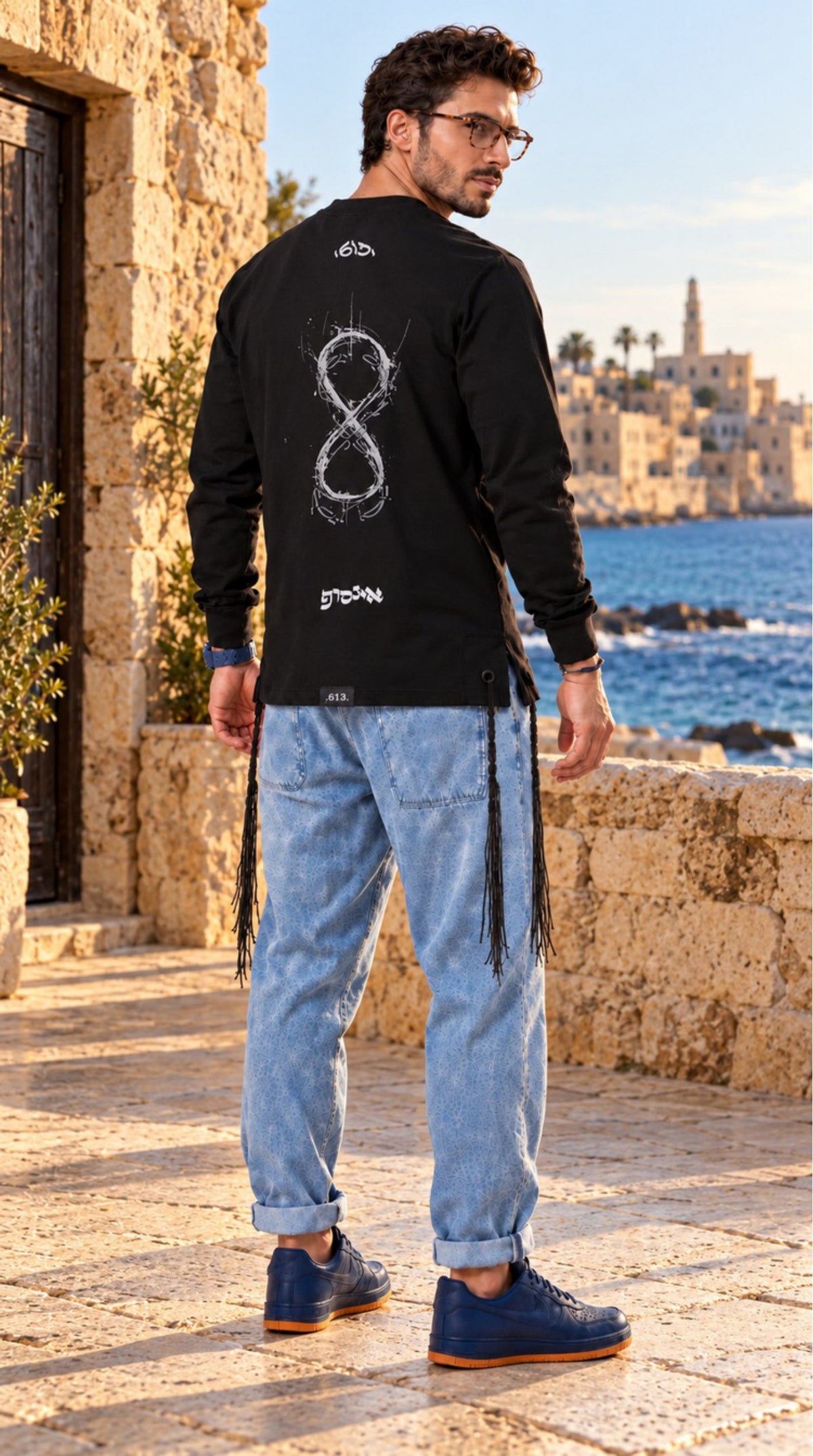 Infinite Presence של 613, חולצת ציצית שחורה עם שרוול ארוך, צילום חוץ לצד מבני אבן והים